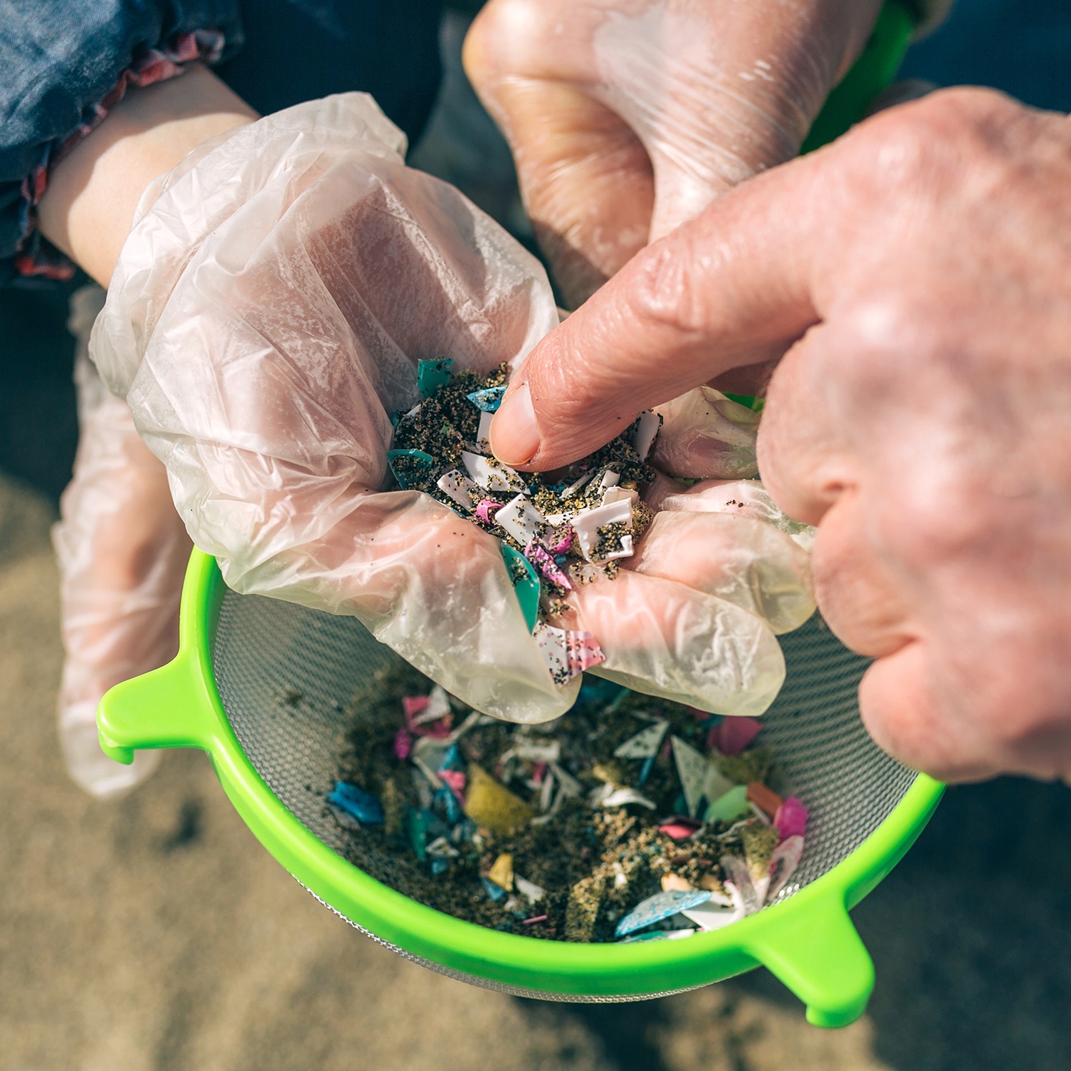 In einer offenen Hand sind kleine Plastikteile und Schmutz zu sehen. Unter der Hand ist ein Sieb, in welchem noch mehr Sand und mehr Plastikteile liegen.